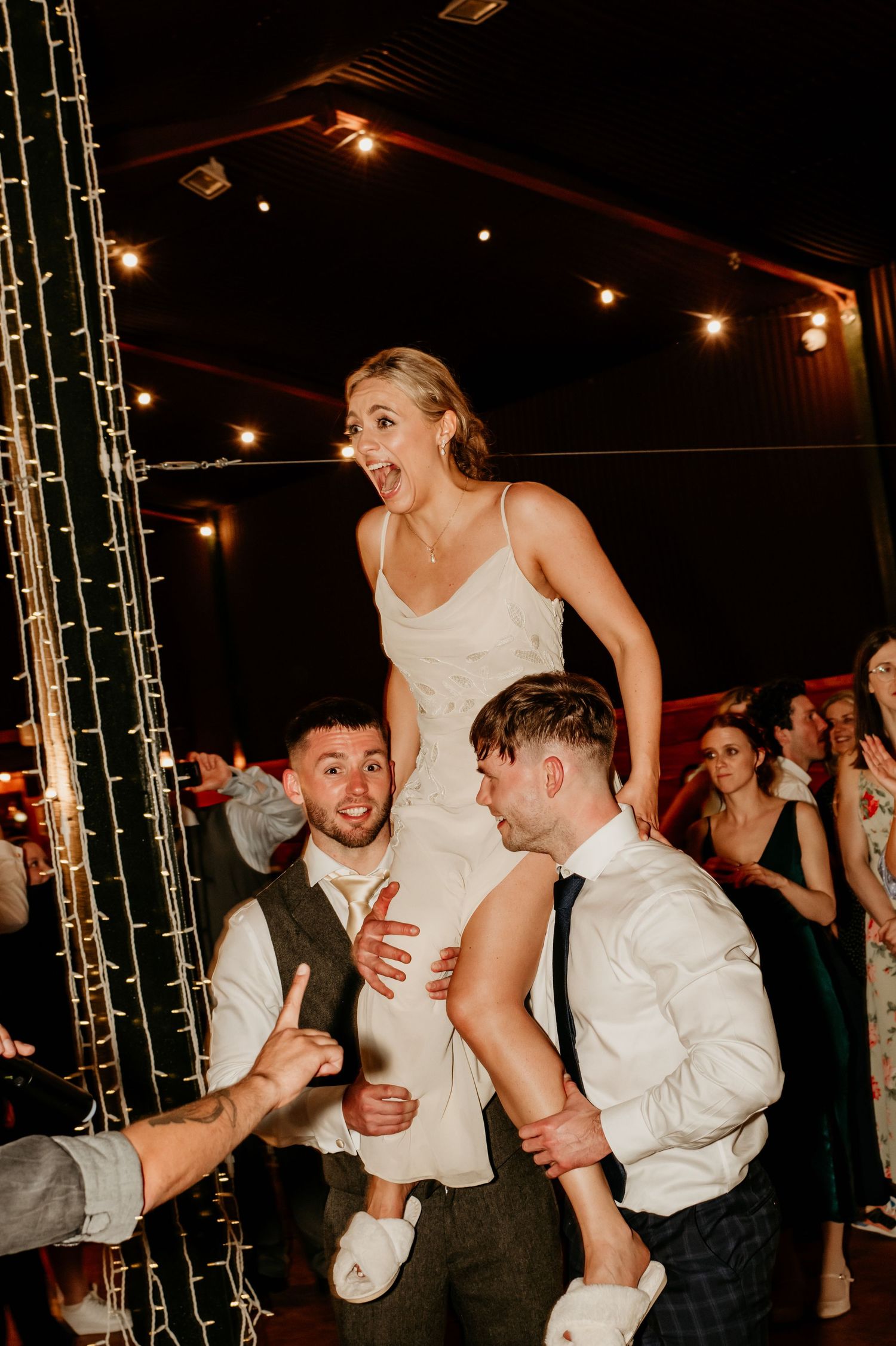 Bride in white dress lifted by groomsmen on wedding dance floor with fairy lights in background.