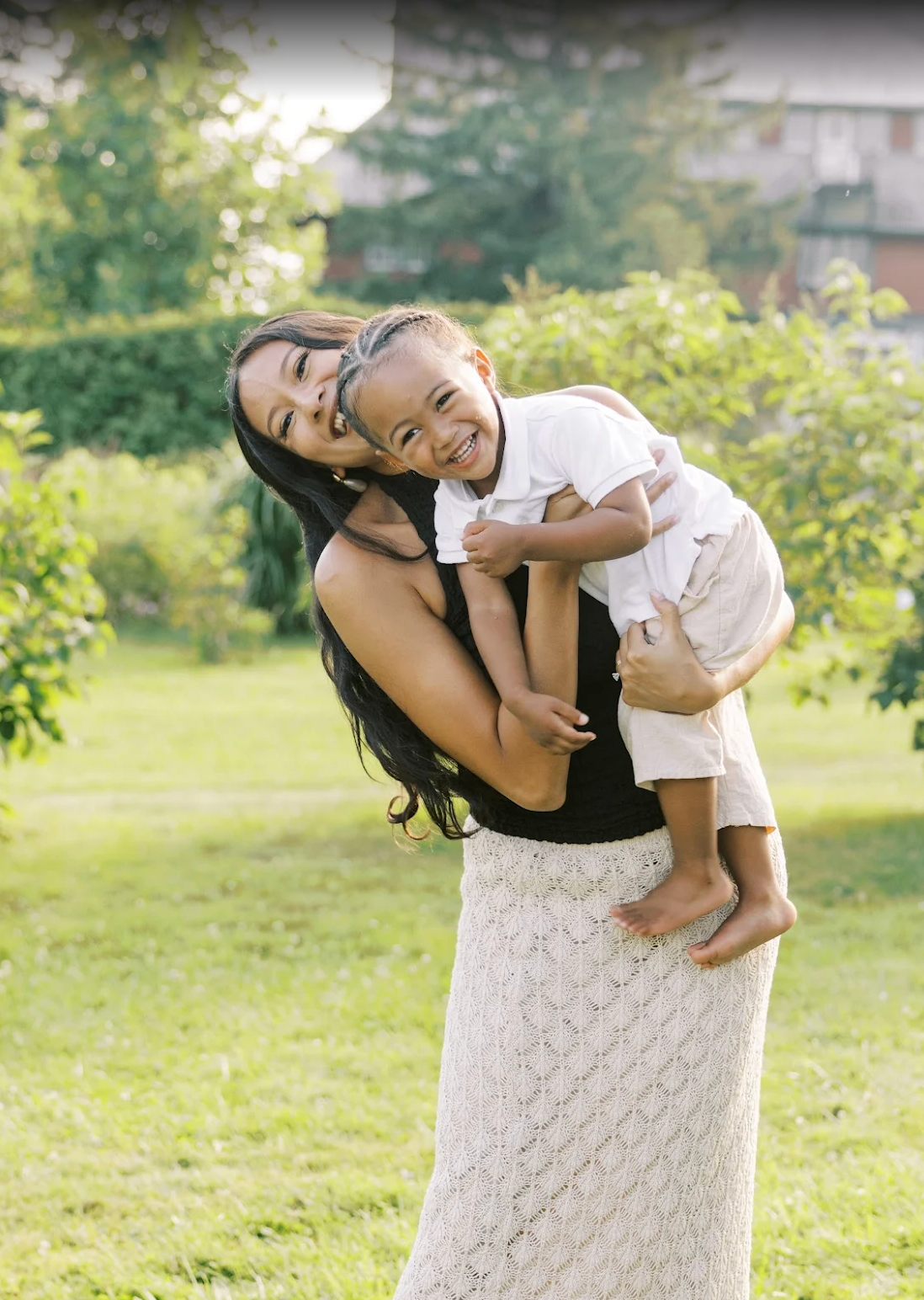A joyful mother holds her laughing young child outdoors in a lush green garden, both smiling brightly.