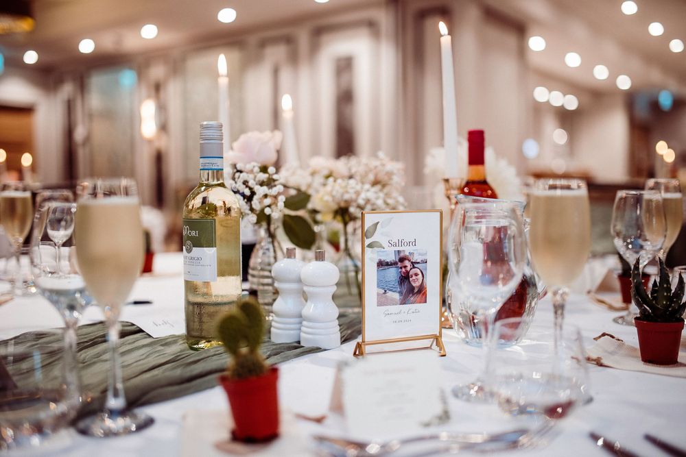 Elegant wedding reception table setting with champagne flutes, white flowers, wine bottles, and a gold framed table number card.