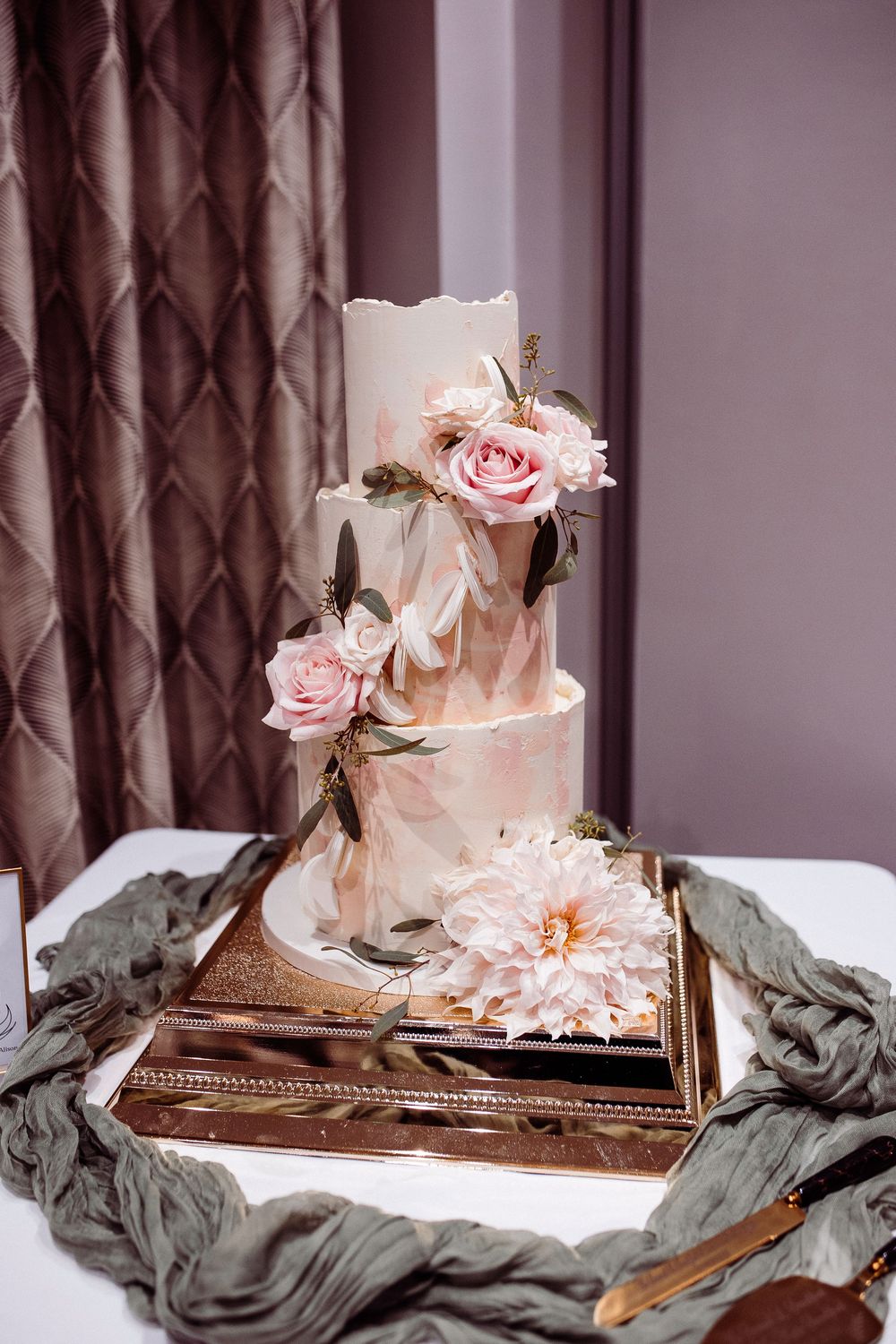 Elegant three-tier white wedding cake adorned with pink roses and dahlias on a gold mirrored tray.