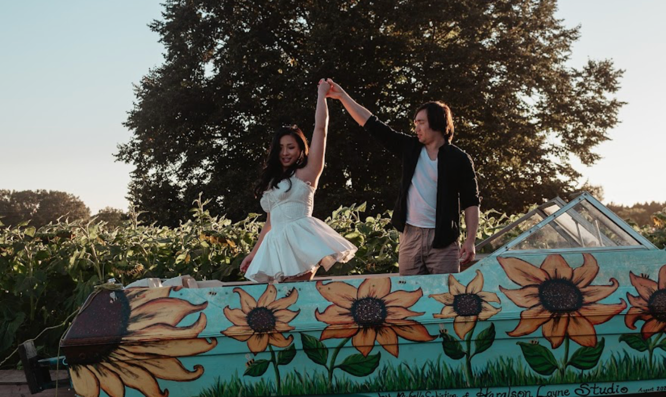 Couple dancing on a colorful sunflower-painted boat outdoors near lush green trees at golden hour.