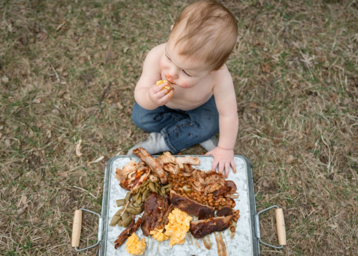 Shirtless toddler sitting on grass eating food from a large aluminum tray filled with BBQ meats and sides.