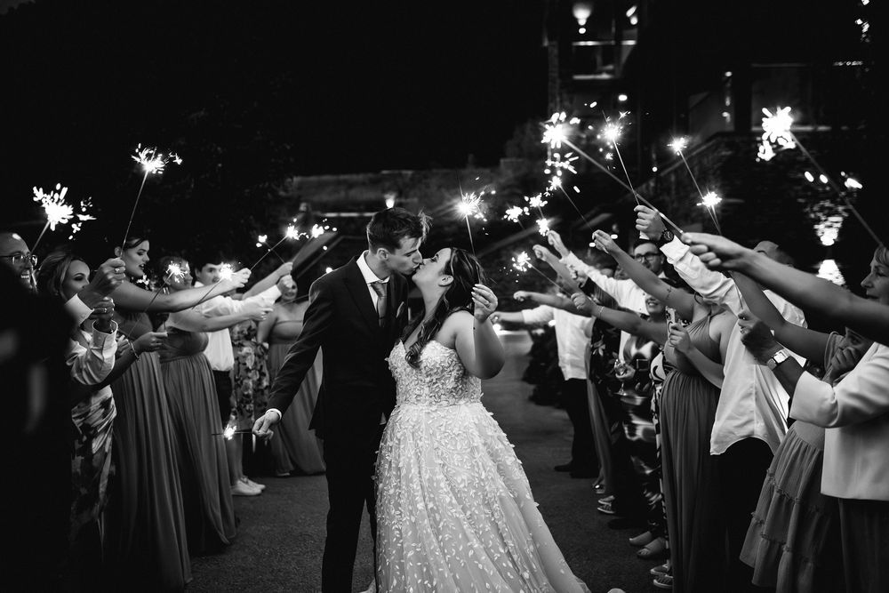 Black and white wedding sparkler exit photo of bride and groom kissing while guests line the path with sparklers.