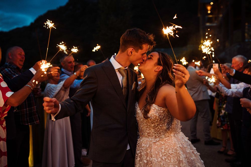 Newlyweds kissing surrounded by guests holding sparklers at a romantic outdoor evening wedding celebration.