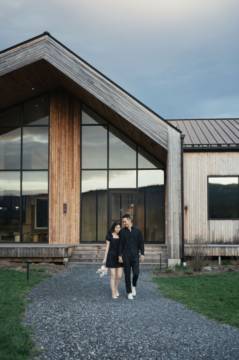 Couple in matching black outfits walking hand-in-hand on gravel path outside modern wooden building at dusk.