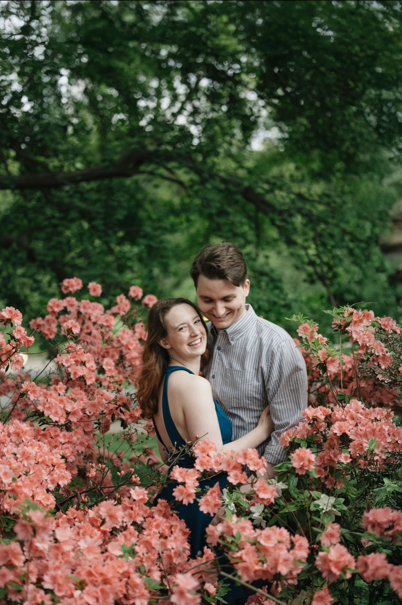 A smiling couple embraces among blooming pink azalea bushes with lush green trees in the background.