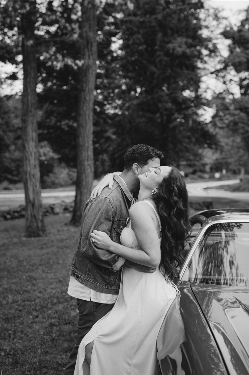 Black and white photo of a couple kissing near a car outdoors, surrounded by tall trees in a romantic setting.