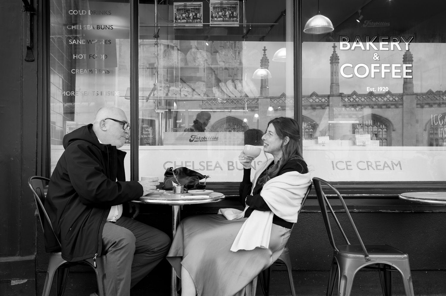 Black and white photo of two people conversing at an outdoor cafe table in front of a bakery and coffee shop.