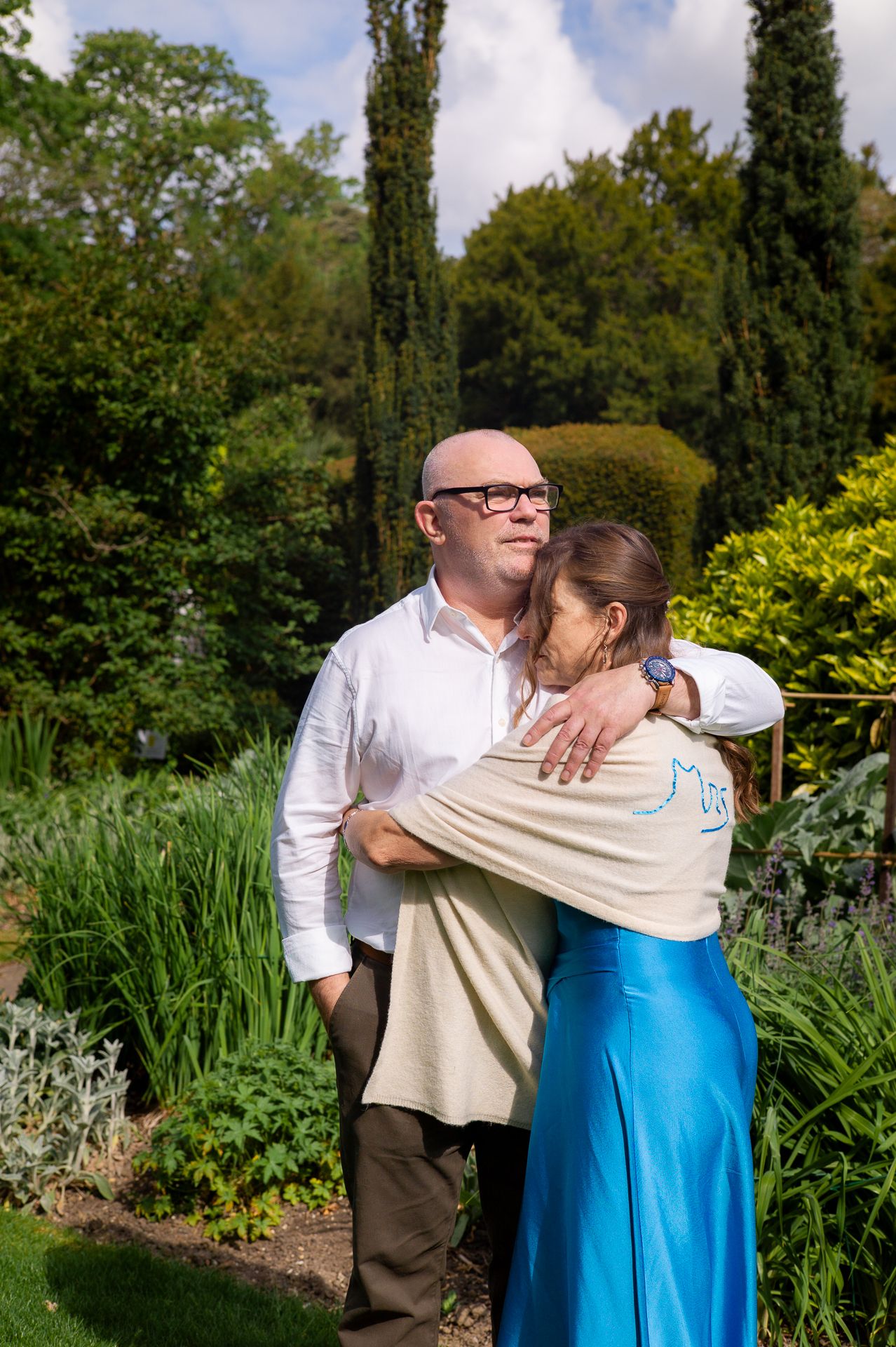 Couple embracing in lush garden, bald man in white shirt hugged by woman in blue skirt and beige top.