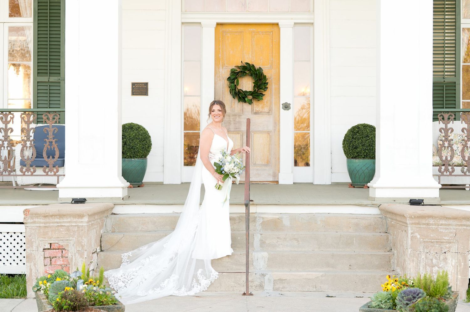 Bride in white gown poses on steps of elegant white Southern mansion with yellow door and wreath.