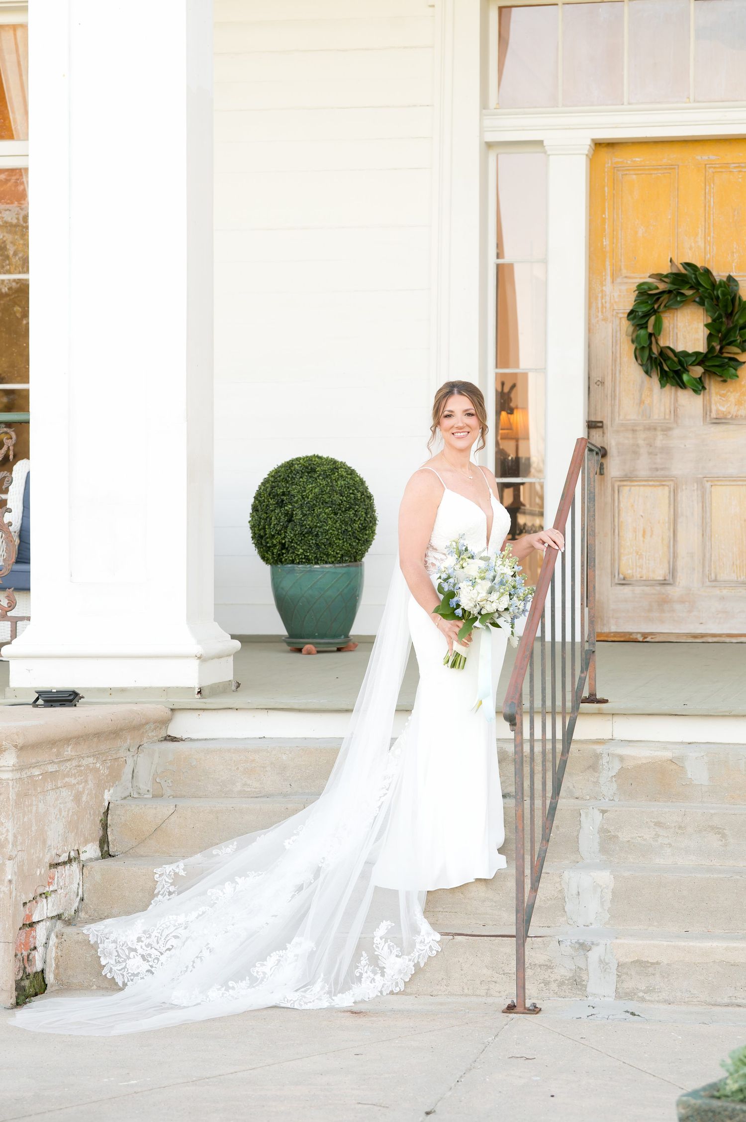 Bride in white lace gown with veil holding bouquet on stone steps outside elegant venue with wreath door.