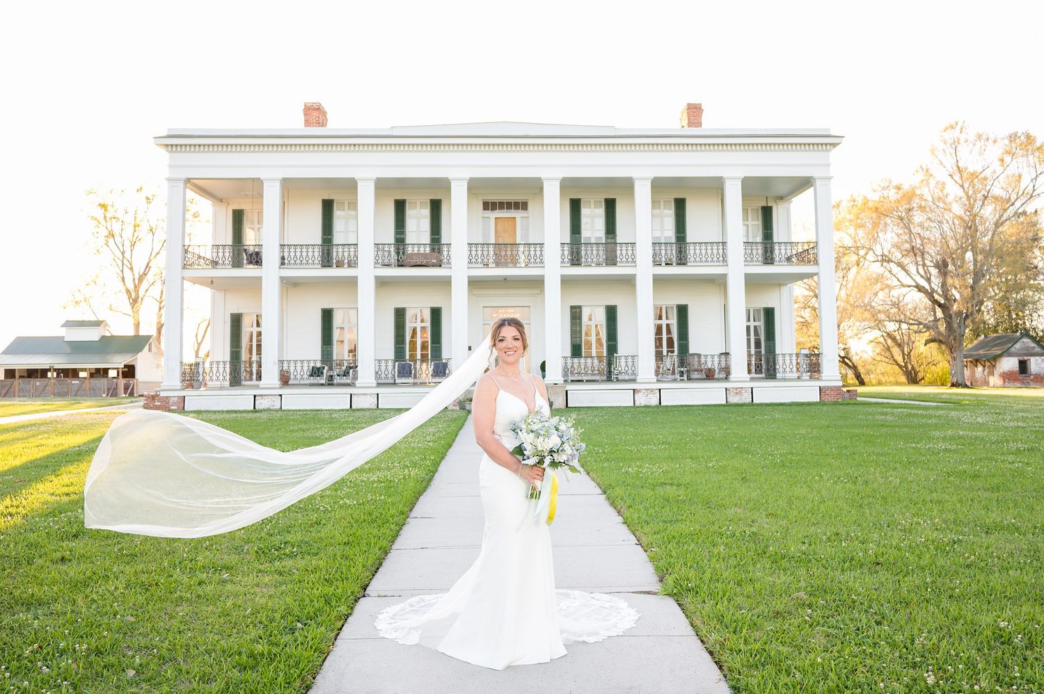 Bride in white gown with flowing veil stands on pathway before grand white Southern plantation wedding venue.