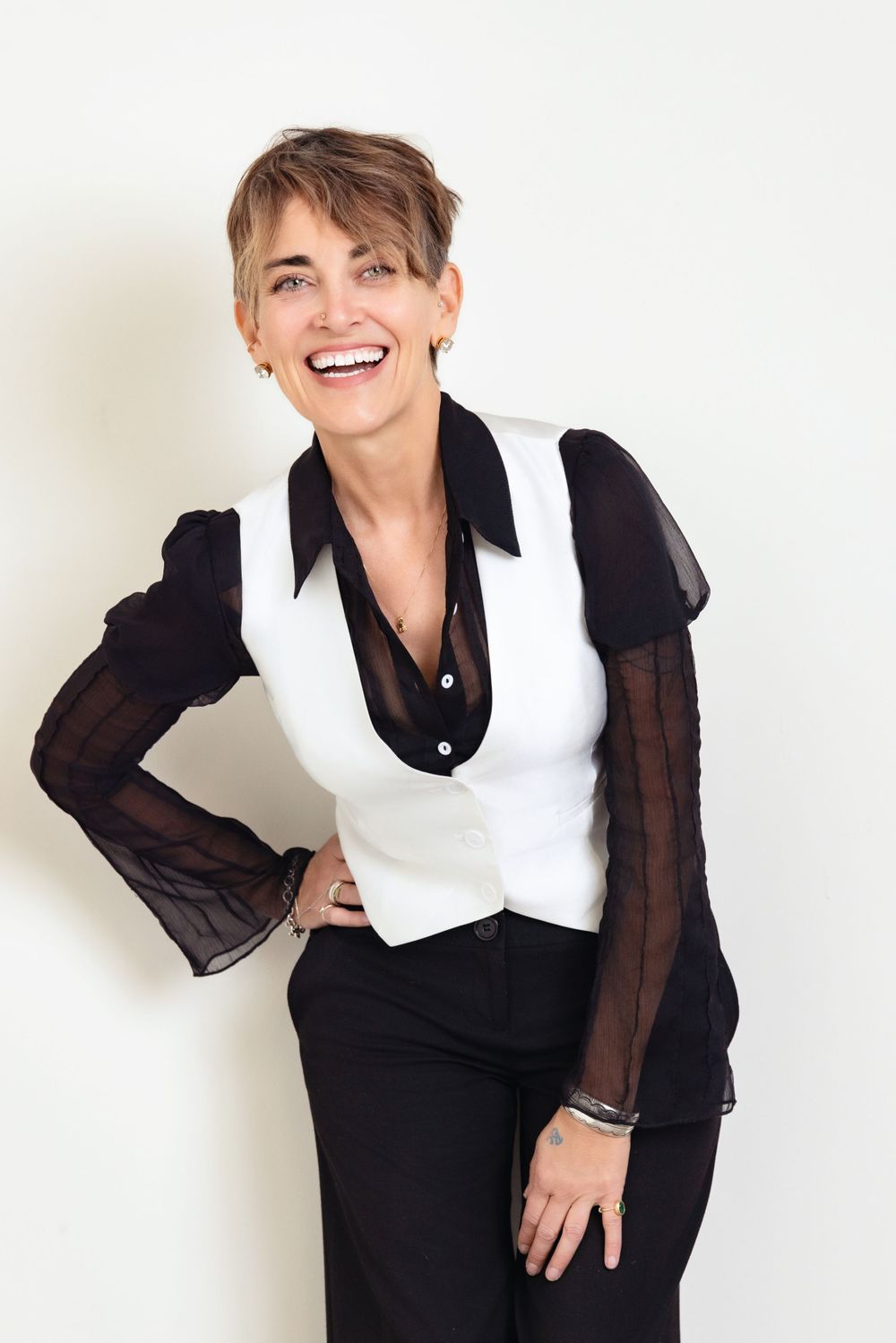 A person wearing a white vest over a black sheer blouse stands against a light wall with a joyful expression.