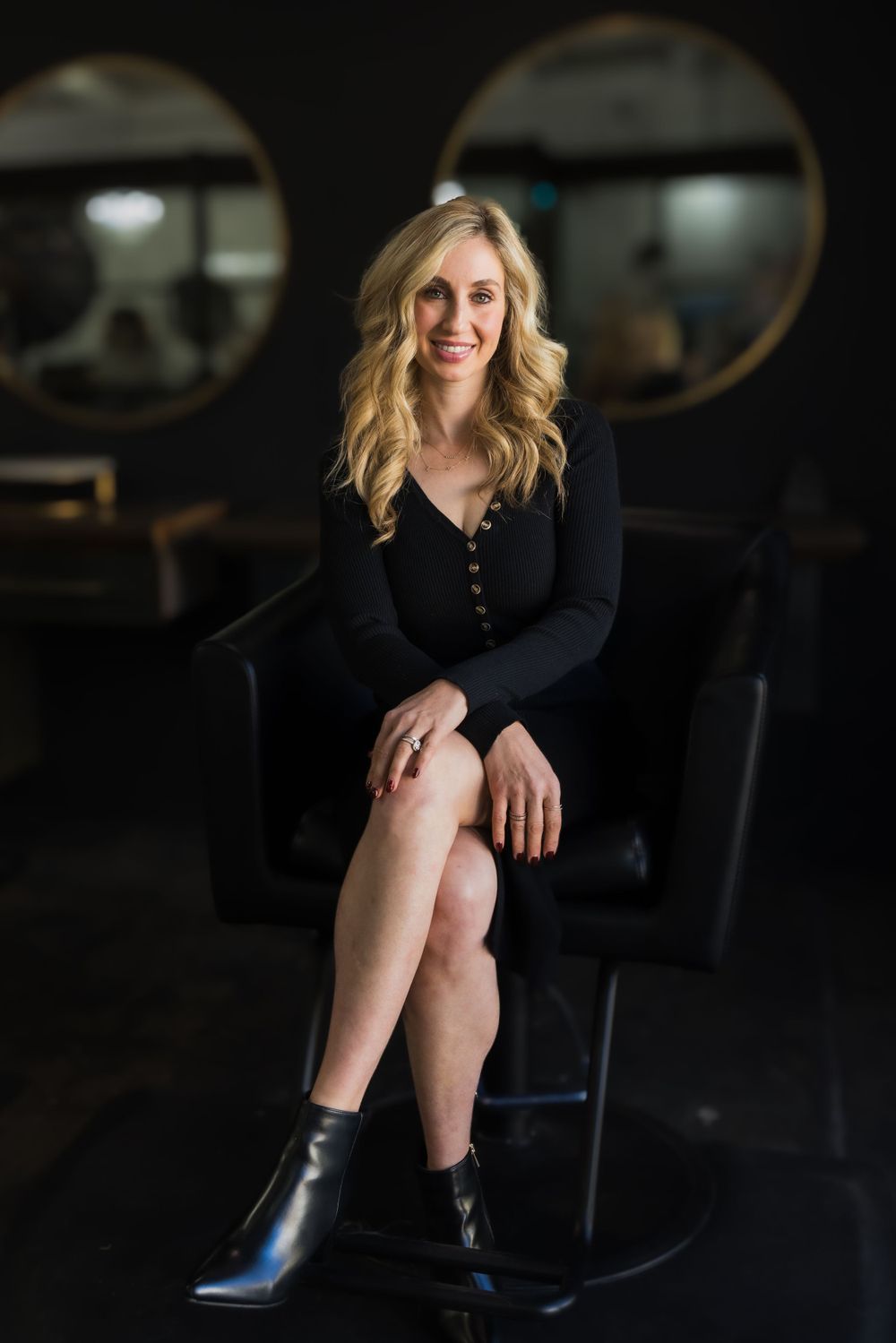 Portrait of a woman with blonde wavy hair wearing a black dress and boots seated in a dark salon setting.
