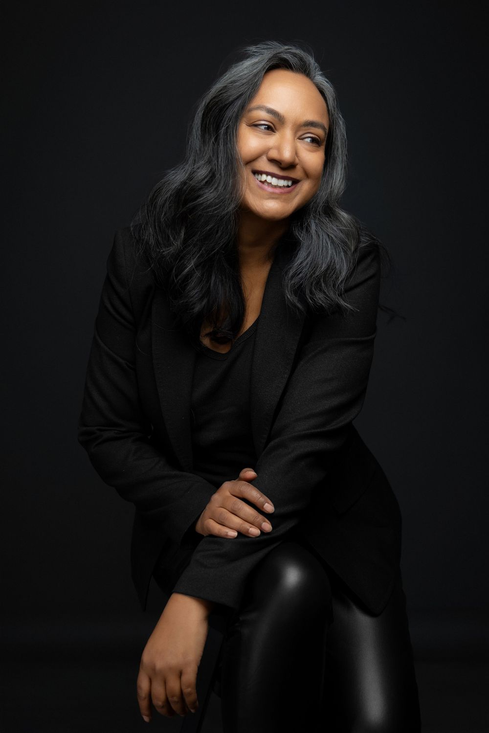 Professional studio portrait of a smiling person with long dark hair wearing a black blazer against a black background.