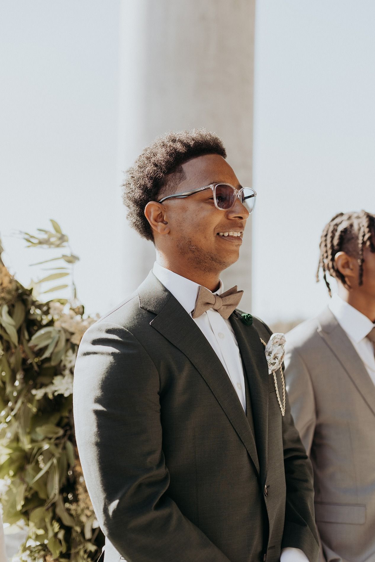 Smiling groom in green suit, bow tie, and sunglasses standing at outdoor wedding ceremony with boutonniere at The Lane.