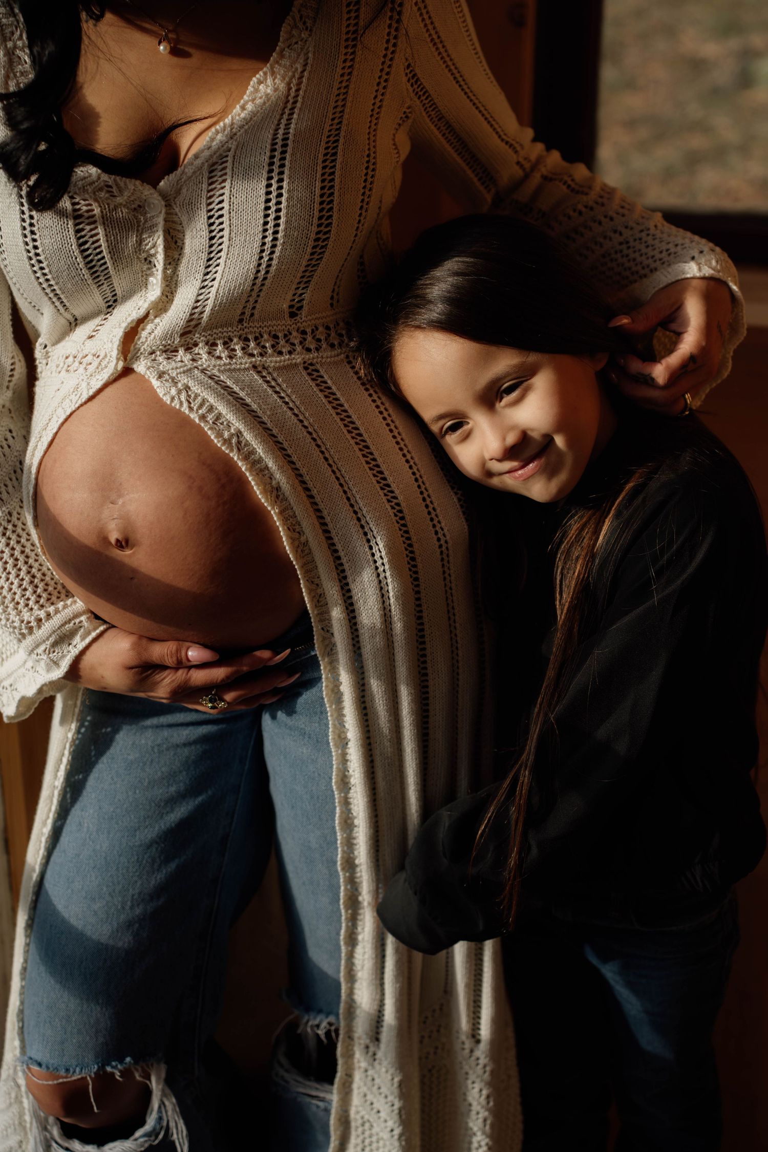 A young girl smiles while leaning against her pregnant mother's belly in a warm, intimate maternity photo.