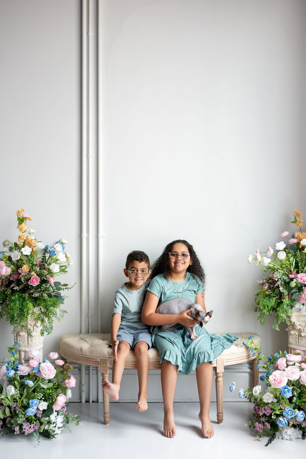 Two children in teal outfits sit on bench holding Sphynx cat with colorful flowers on both sides.
