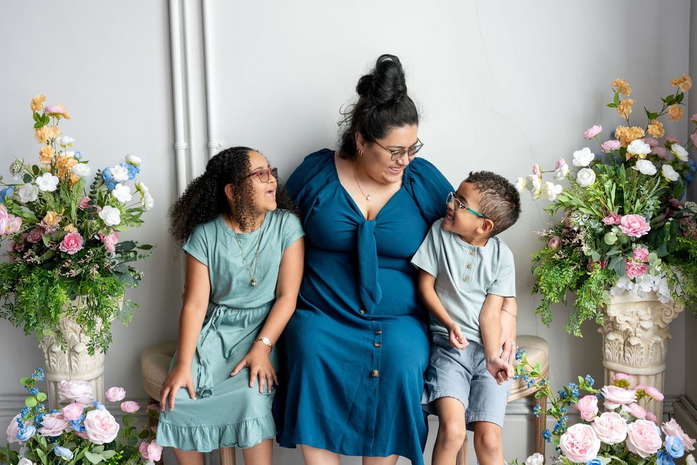 A mother and her two children share a candid moment on a bench surrounded by colorful spring floral arrangements.