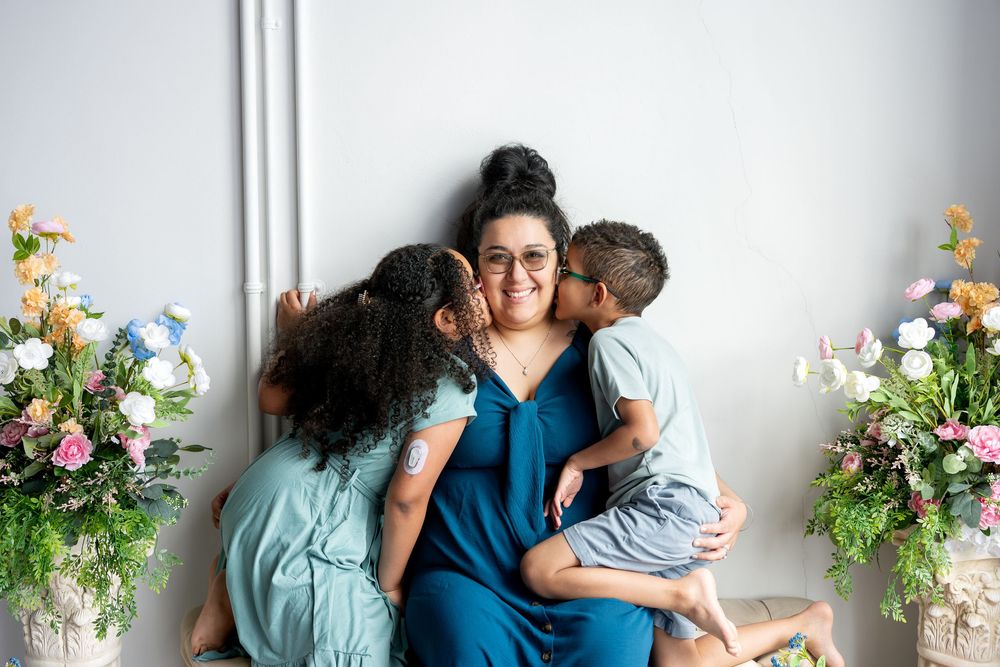 Two children lovingly kiss their smiling mother on the cheeks in a bright studio with floral decor.