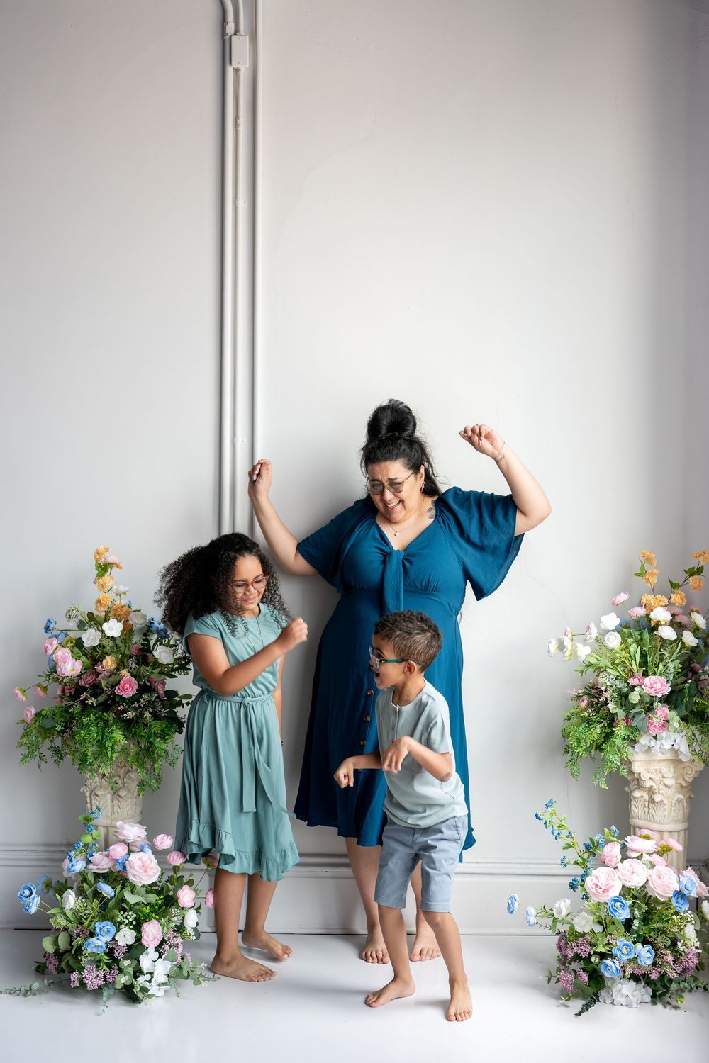 A mother and her two children dance joyfully in a bright studio decorated with colorful spring floral arrangements.