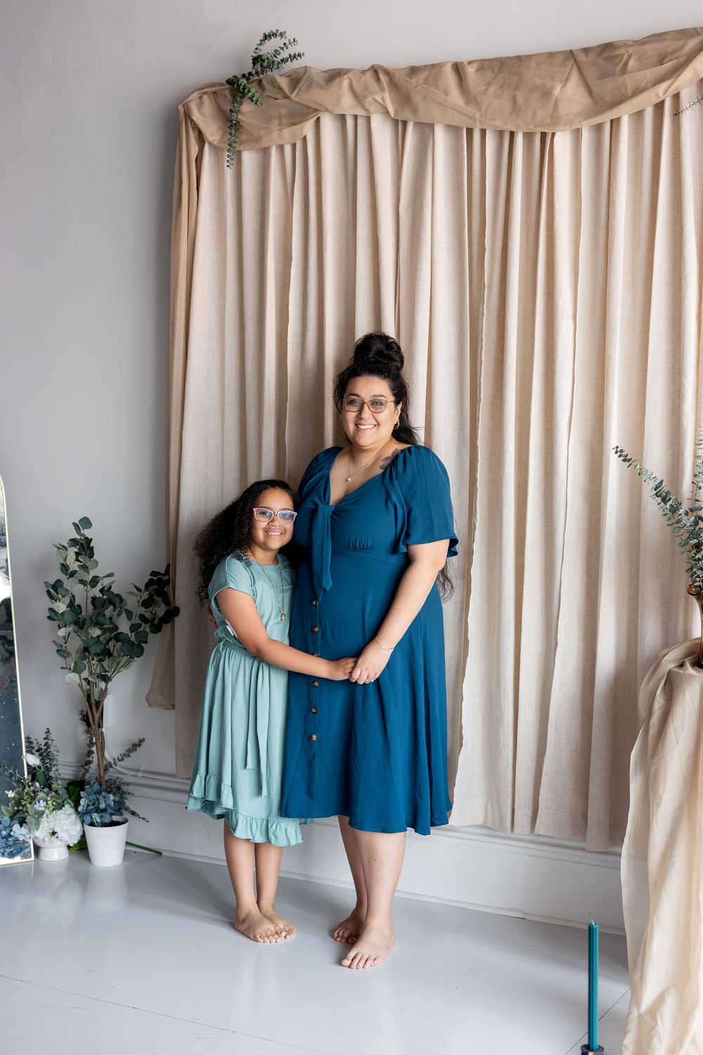 A girl in a mint dress stands beside a pregnant woman in a teal dress in a studio.