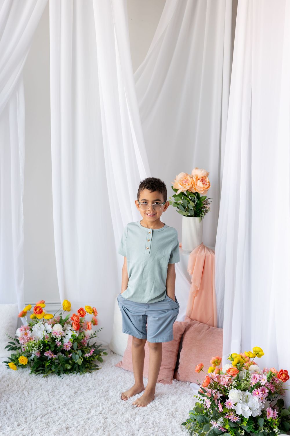 A boy in a mint shirt and gray shorts stands among colorful floral arrangements in a studio.
