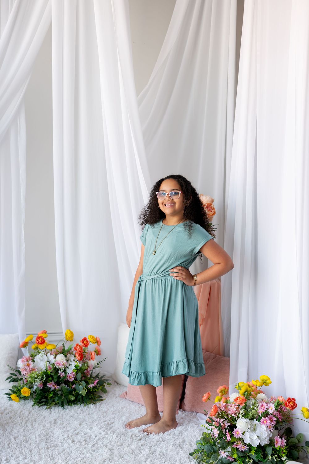 A girl in a teal dress and glasses poses barefoot among colorful flowers in a white draped studio.