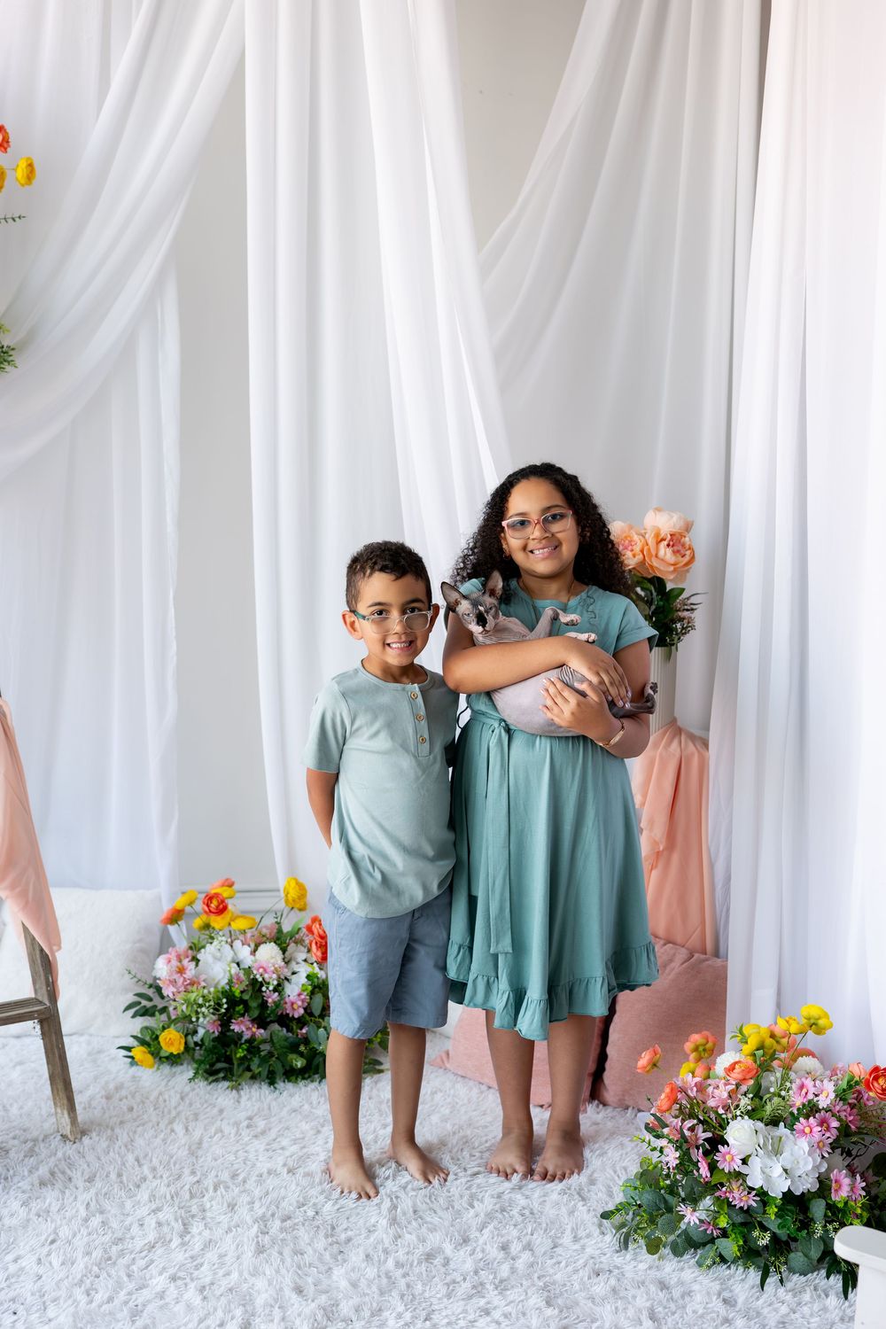 A boy and girl wearing glasses and teal outfits smile together in a bright floral studio backdrop.