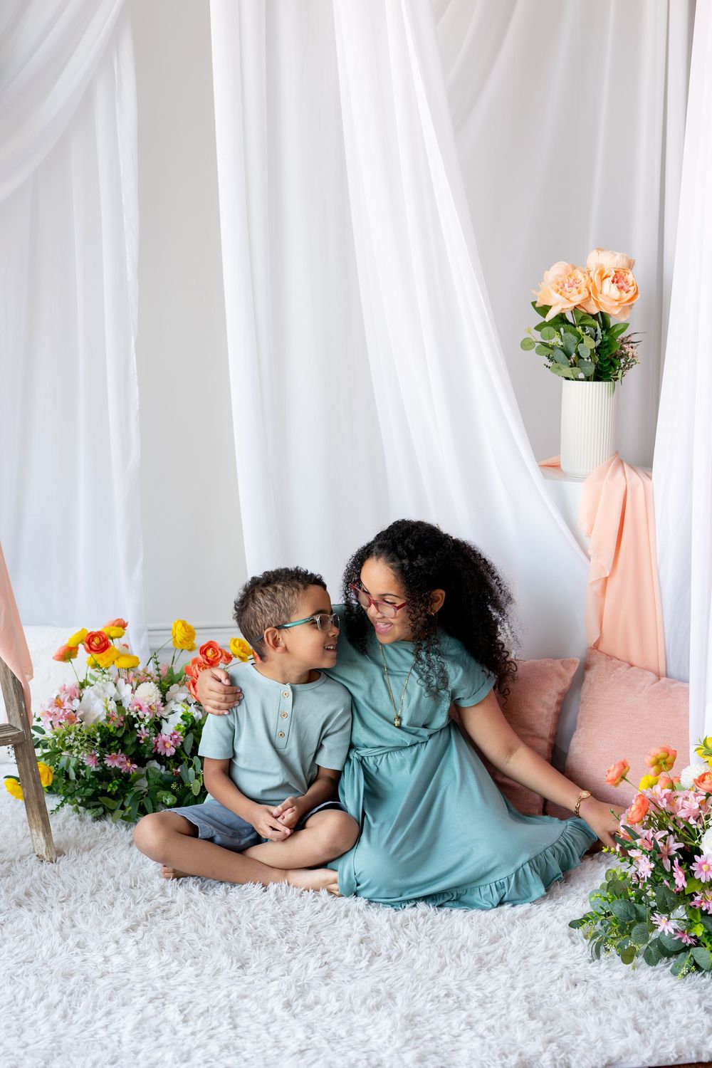A girl in a teal dress hugs a younger boy as they sit together among flowers in a white studio.