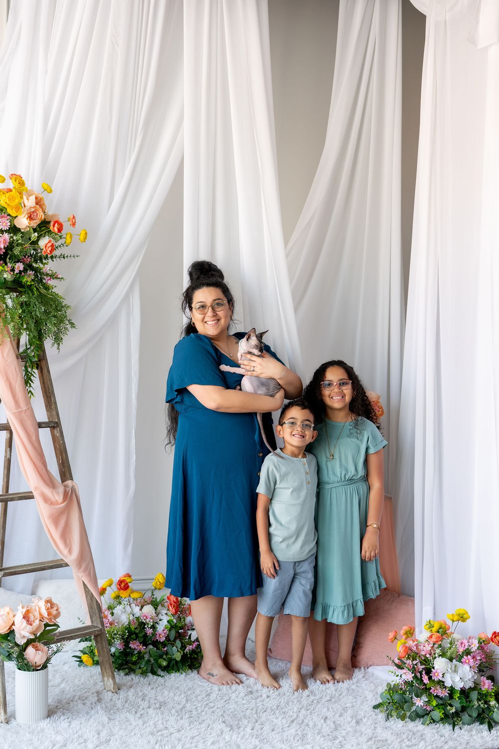 A woman in a blue dress holds a cat while posing with two children in a bright floral studio.
