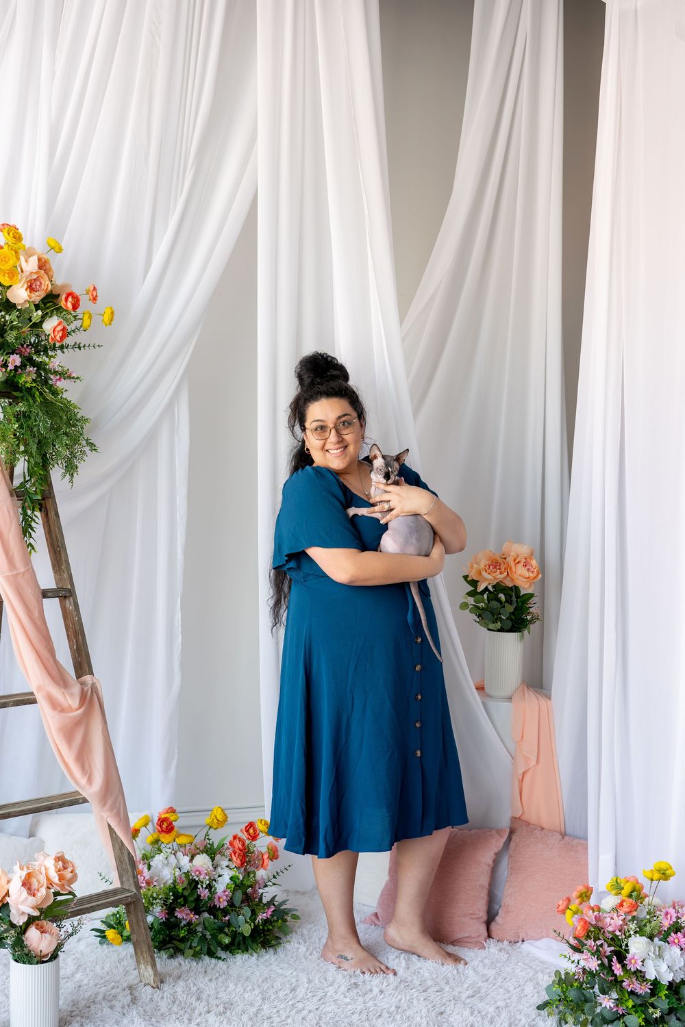 Smiling woman in blue dress holds a Sphynx cat surrounded by colorful flowers in a white studio.
