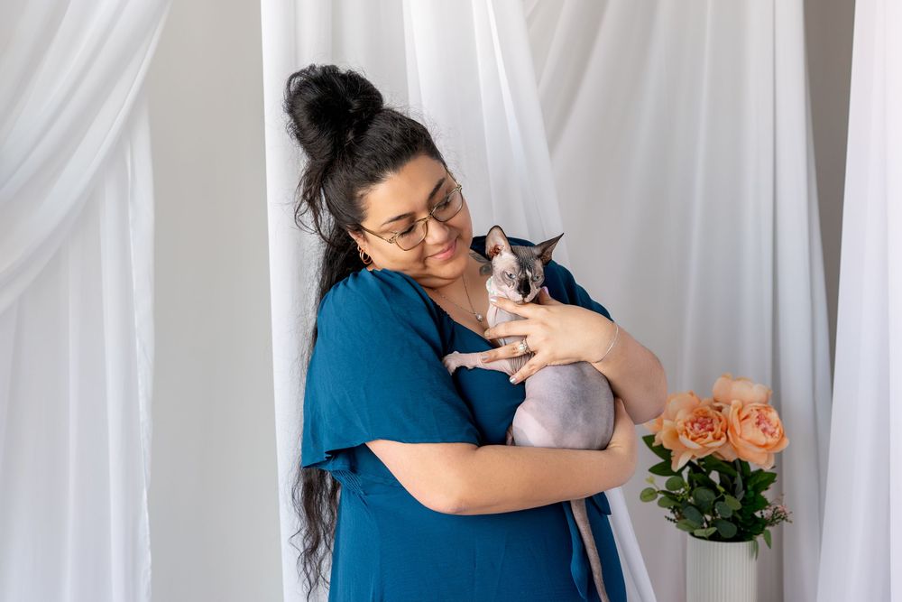 Woman in blue dress tenderly holds a Sphynx cat near white curtains and peach roses.