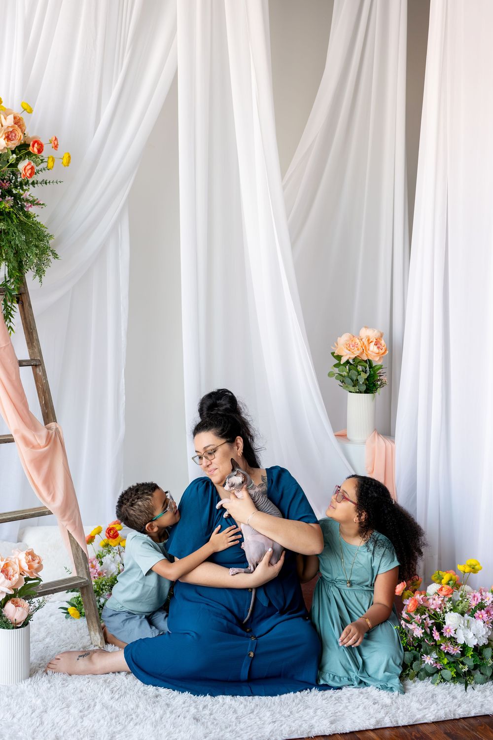 Pregnant woman in blue dress sits with two children and a cat amid flowers and white curtains.