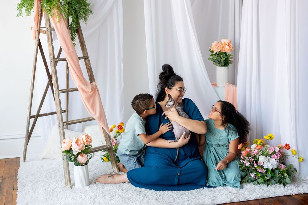 A mother and her two children share a tender moment with a cat in a floral studio setting.