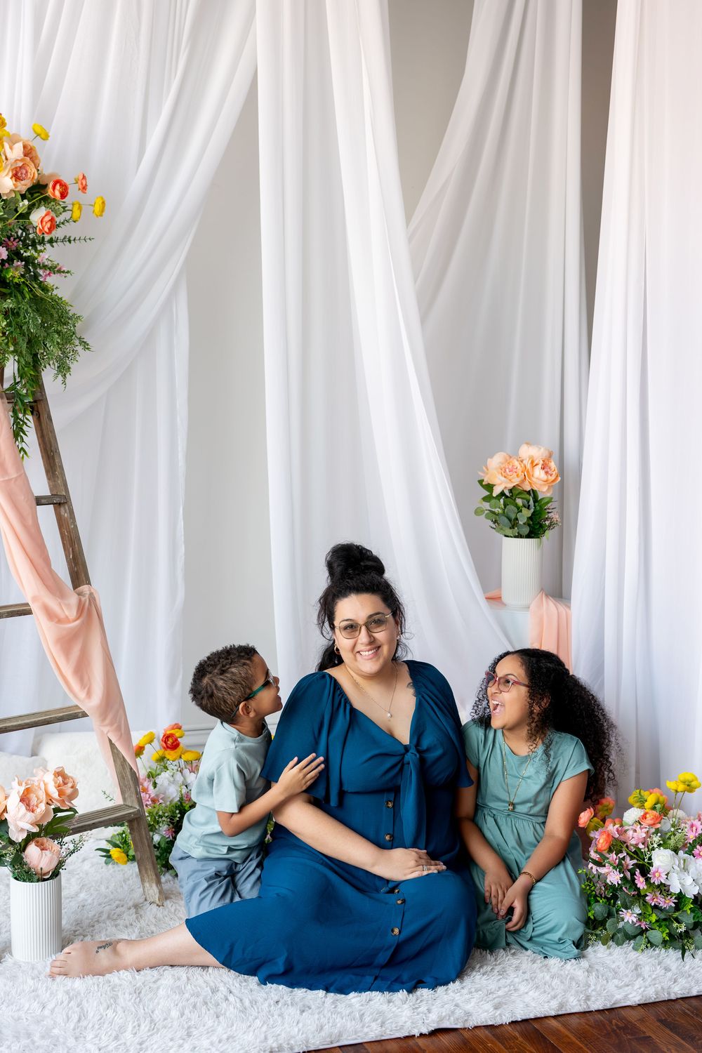 A pregnant mother laughs with her two children in a white studio decorated with spring flowers.