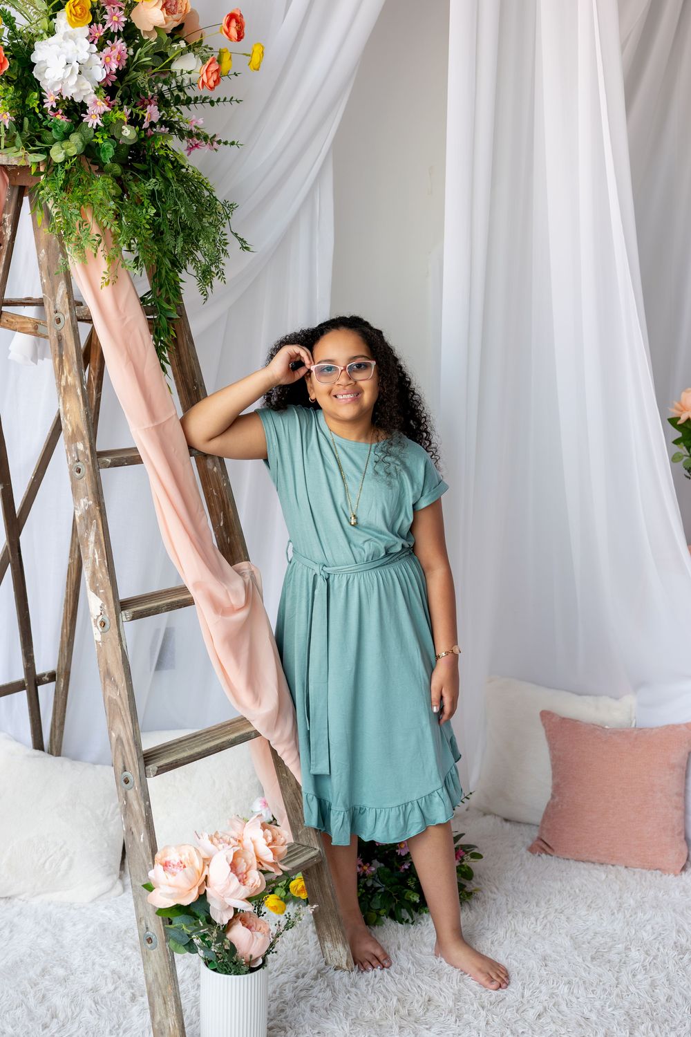 A young girl in a teal dress poses by a floral ladder in a bright white studio.