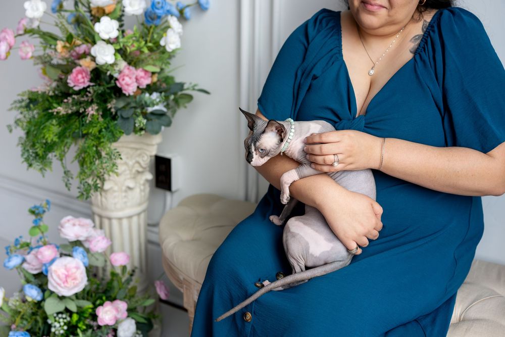 Close-up of woman in teal dress holding a Sphynx cat beside pink and blue floral arrangement.