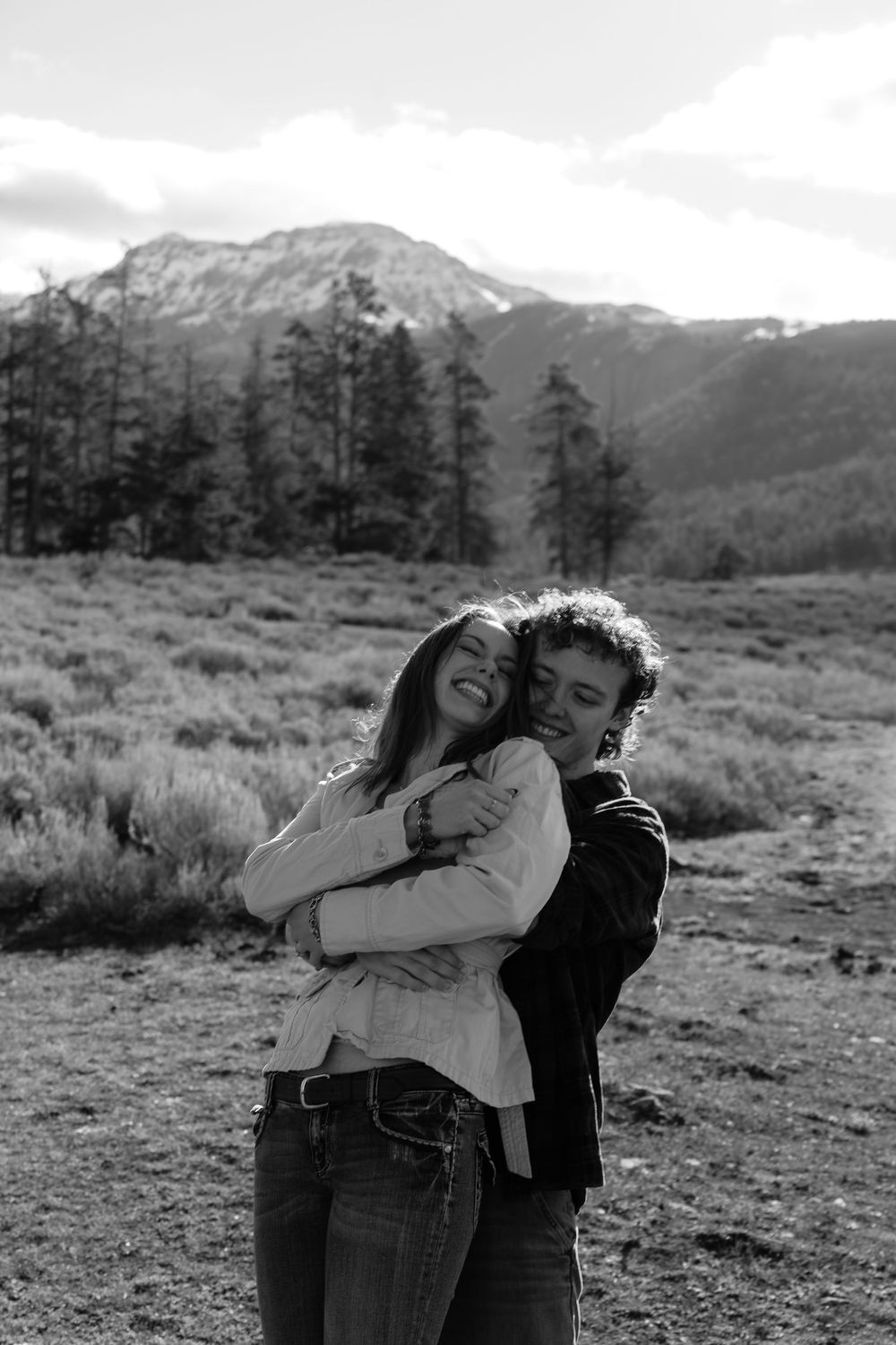 Black and white photo of a happy couple embracing and smiling in a mountain meadow with snow-capped peaks behind them.