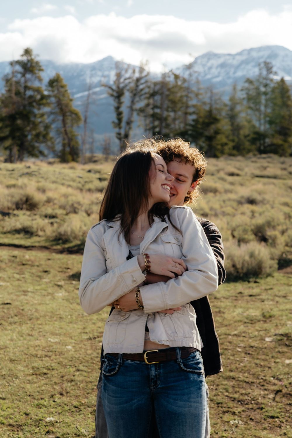 Couple embracing in a mountain meadow, woman in white jacket smiling as man kisses her cheek.