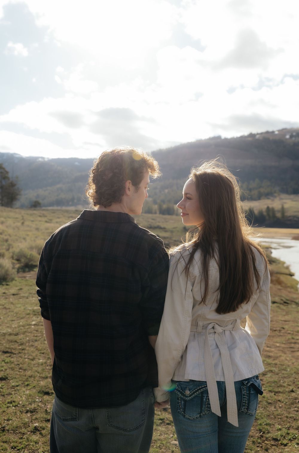 Couple holding hands while looking at each other in a sunlit mountain landscape with river views.