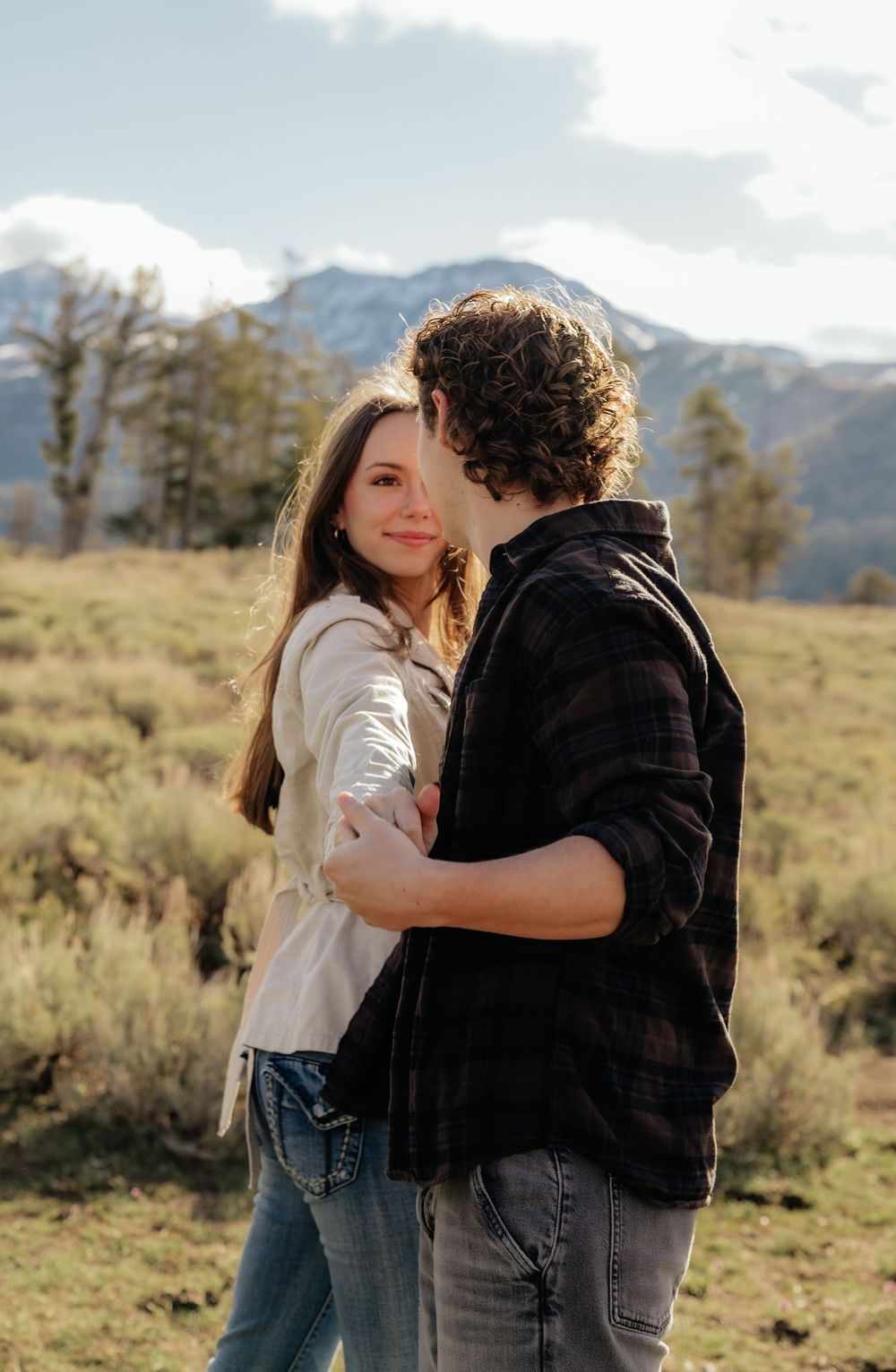 Couple embracing in a mountain meadow, woman smiling at camera wearing cream shirt and jeans.