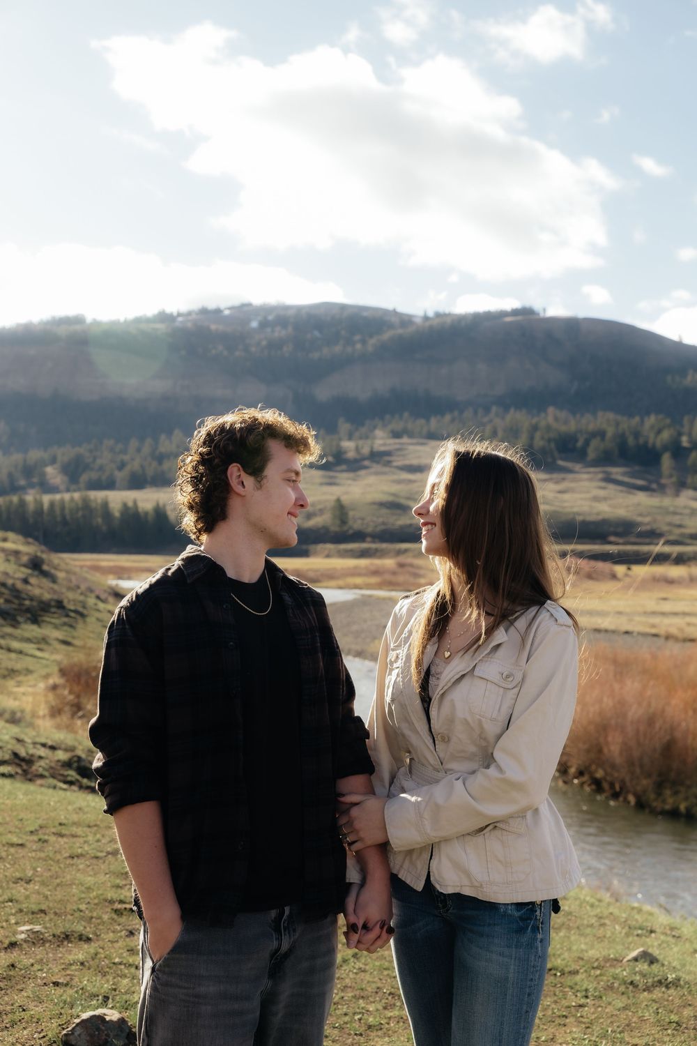 Young couple holding hands and gazing at each other in a scenic mountain valley with a river backdrop.