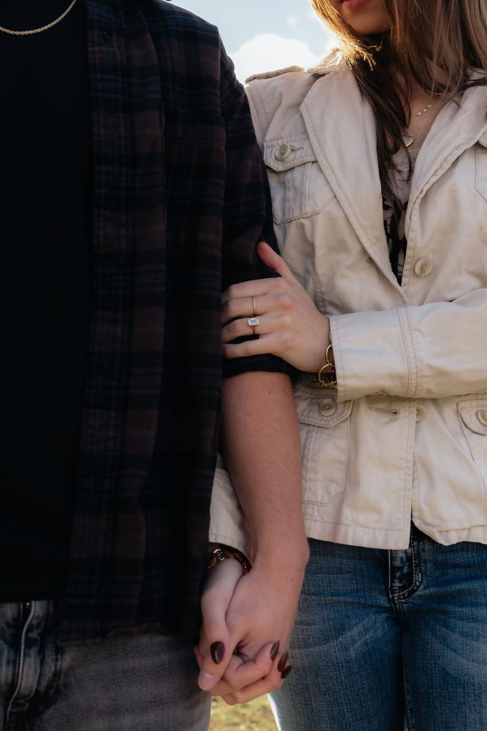 Couple holding hands outdoors, woman wearing engagement ring and beige jacket, man in plaid shirt and jeans.