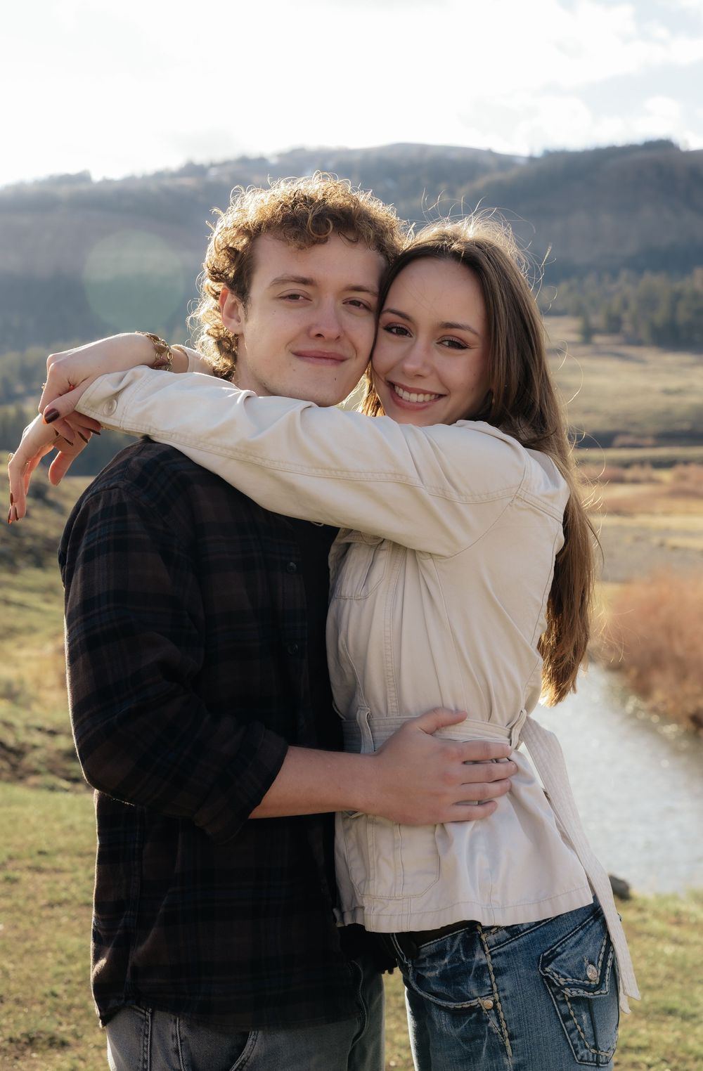 A young couple embraces and smiles in a sunny outdoor mountain valley setting near a winding river.