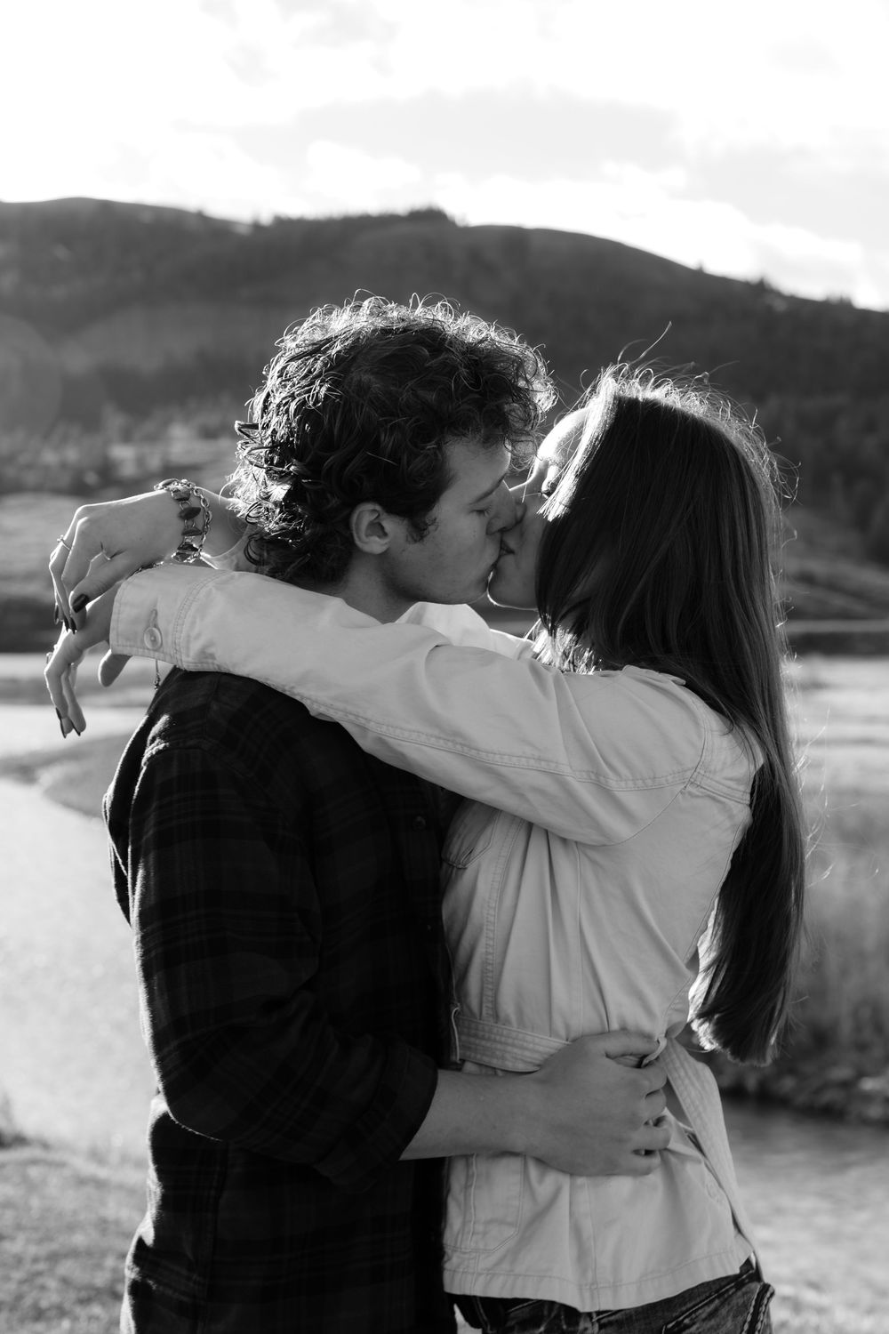 Couple sharing a romantic kiss outdoors with mountain lake backdrop in black and white photo.