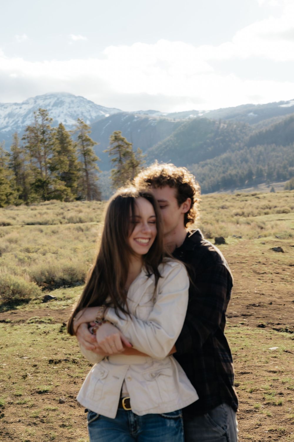 Couple embracing in a mountain meadow with snow-capped peaks, woman smiling joyfully.