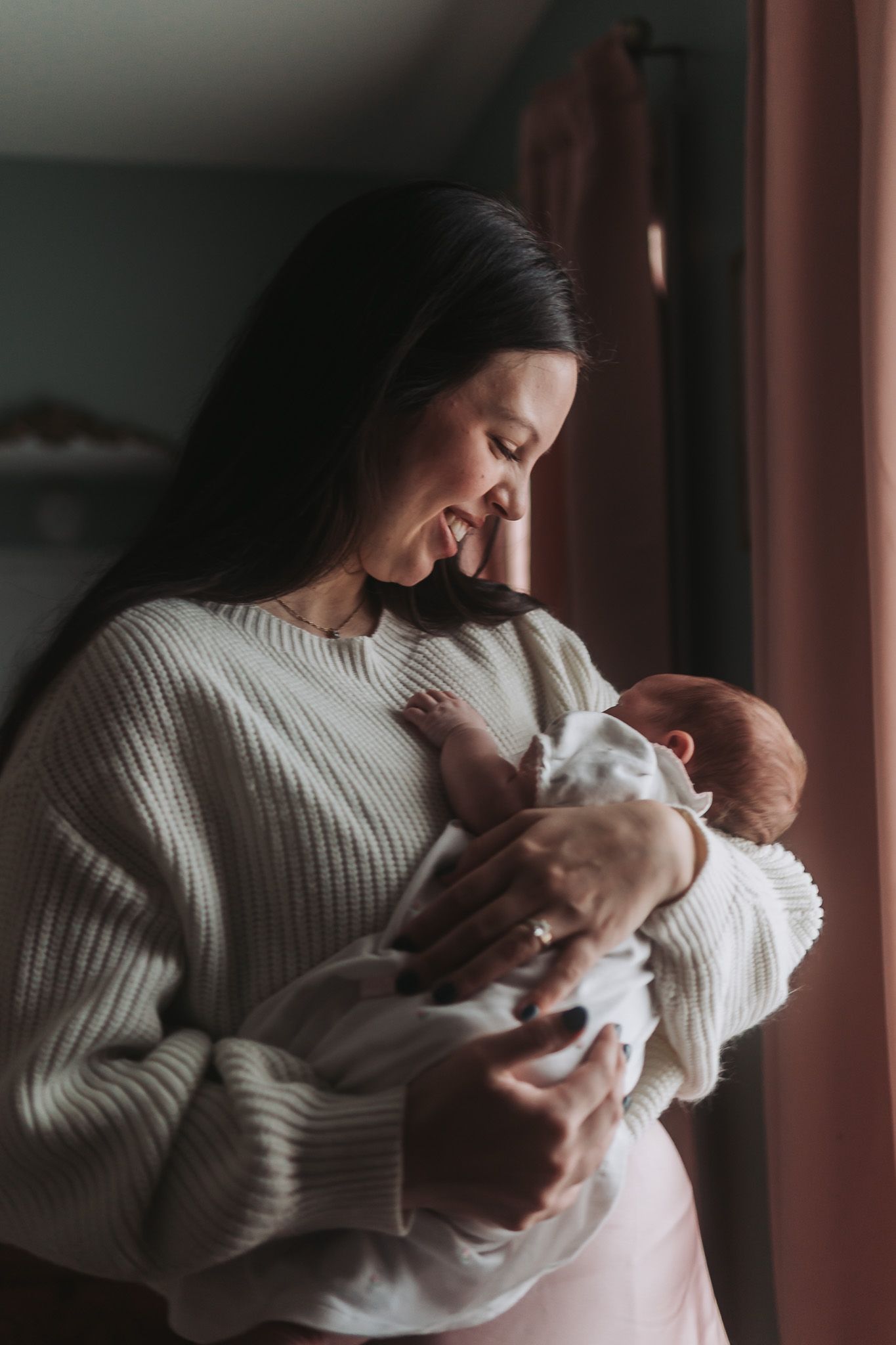 Smiling dark-haired mother in cream sweater tenderly holding newborn baby by window.