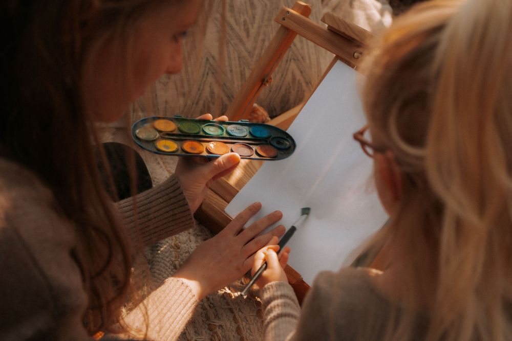 Two girls painting with watercolors on an easel, sharing a colorful paint palette in warm natural light.