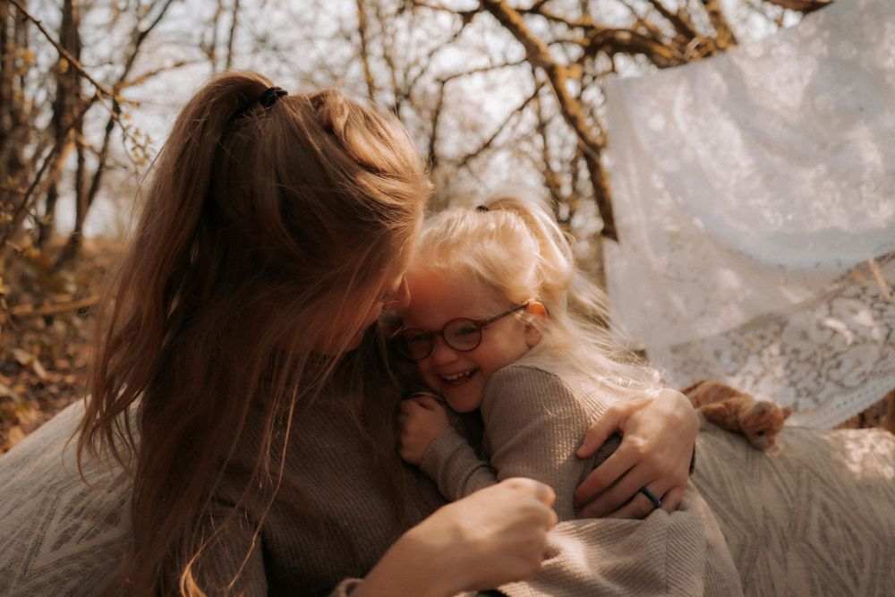 A mother and young girl with glasses share a warm embrace outdoors among autumn trees.